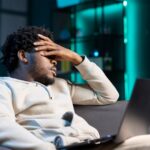 Stellar Photo Recovery Review TrueTech. Man suffering from headache and frustration, sitting on couch at home, after accidentally deleting all his photos from the laptop on his lap.