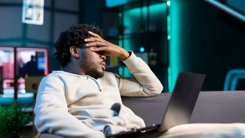 Stellar Photo Recovery Review TrueTech. Man suffering from headache and frustration, sitting on couch at home, after accidentally deleting all his photos from the laptop on his lap.