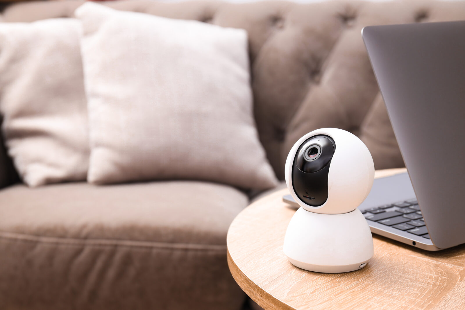 White indoor smart security camera on a side table beside a sofa and laptop